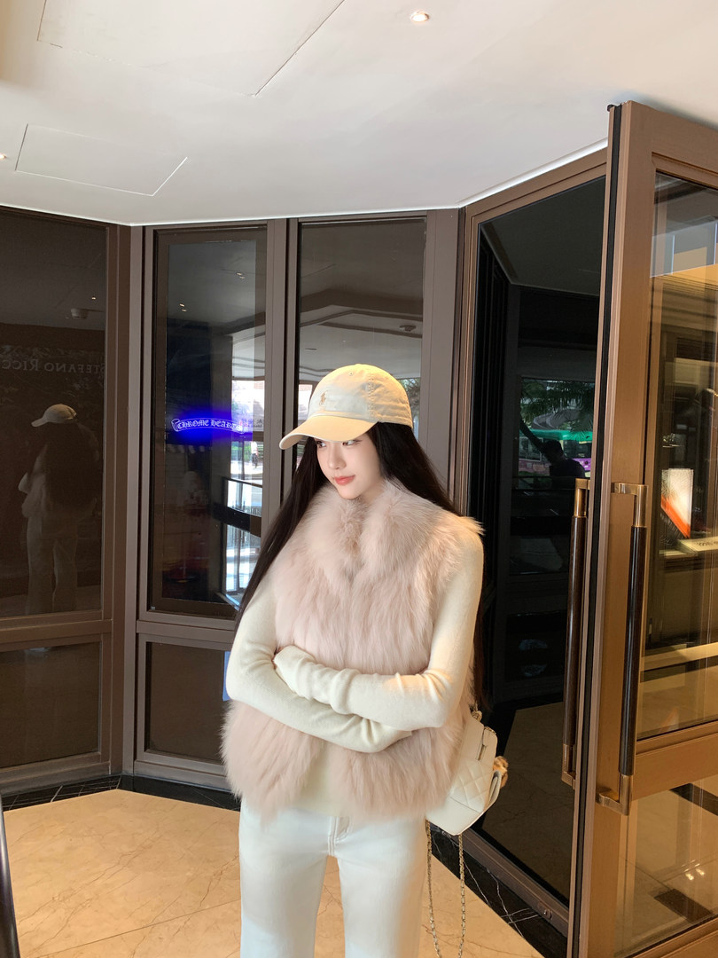 Woman wearing a light pink faux fur vest over a cream sweater, paired with white pants and a beige baseball cap, standing indoors near glass doors.