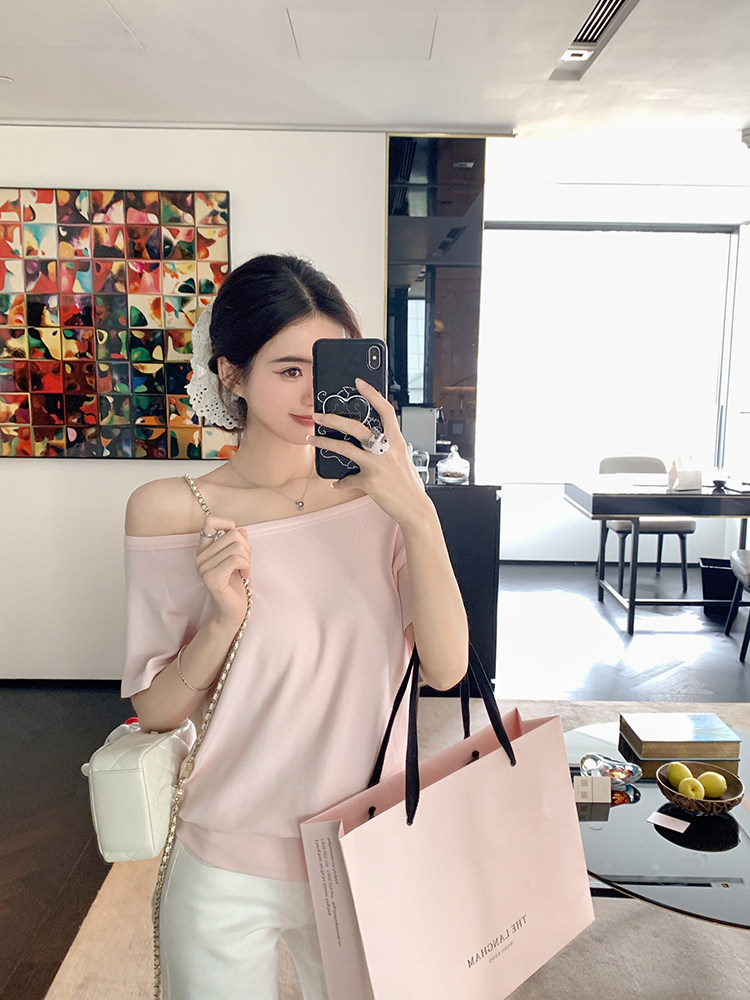 Woman wearing a light pink off-shoulder top, holding a phone and shopping bag in a modern interior.