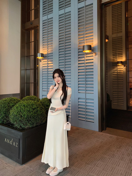 Woman wearing a white dress with a small white handbag, standing in front of a building with shutters.