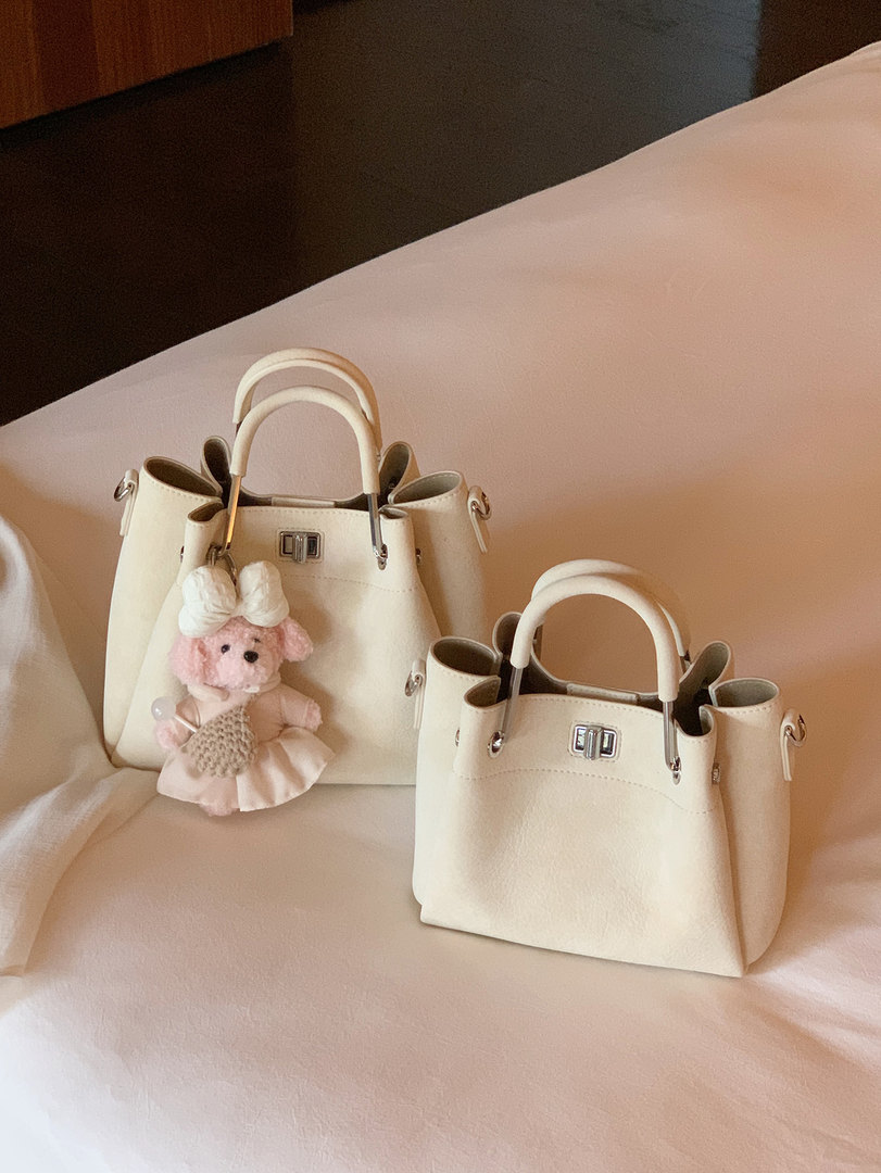 Two cream-colored handbags with structured handles and a pink teddy charm displayed on a bed