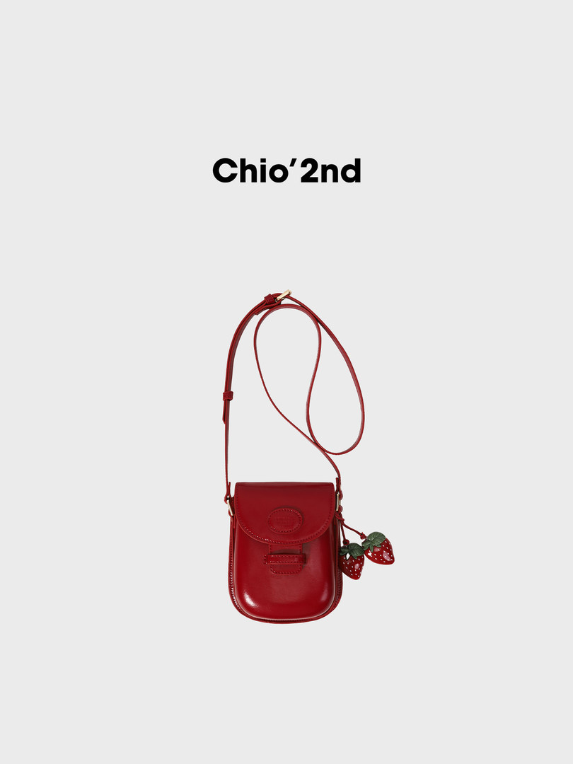 Red leather mini crossbody bag with strawberry charm on a plain background