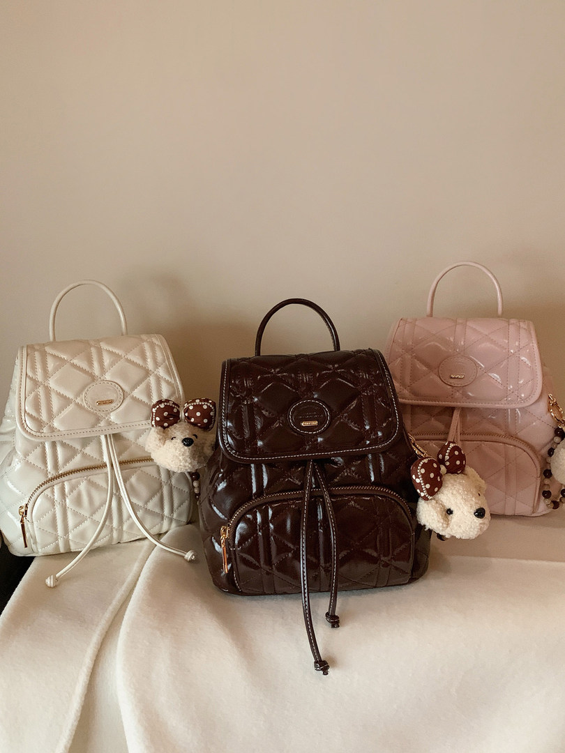 Three quilted backpacks in white, brown, and pink with teddy bear keychains displayed on a bed