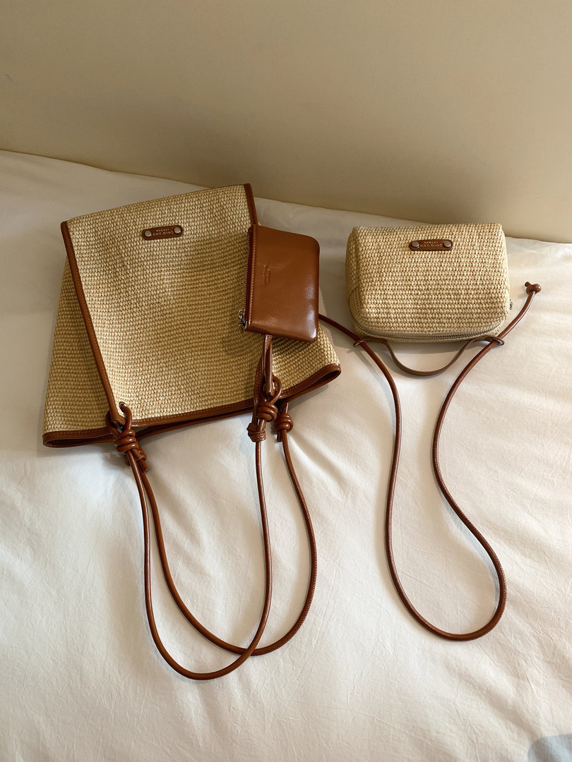 Two woven tan crossbody bags with brown leather trim and long straps on a bed