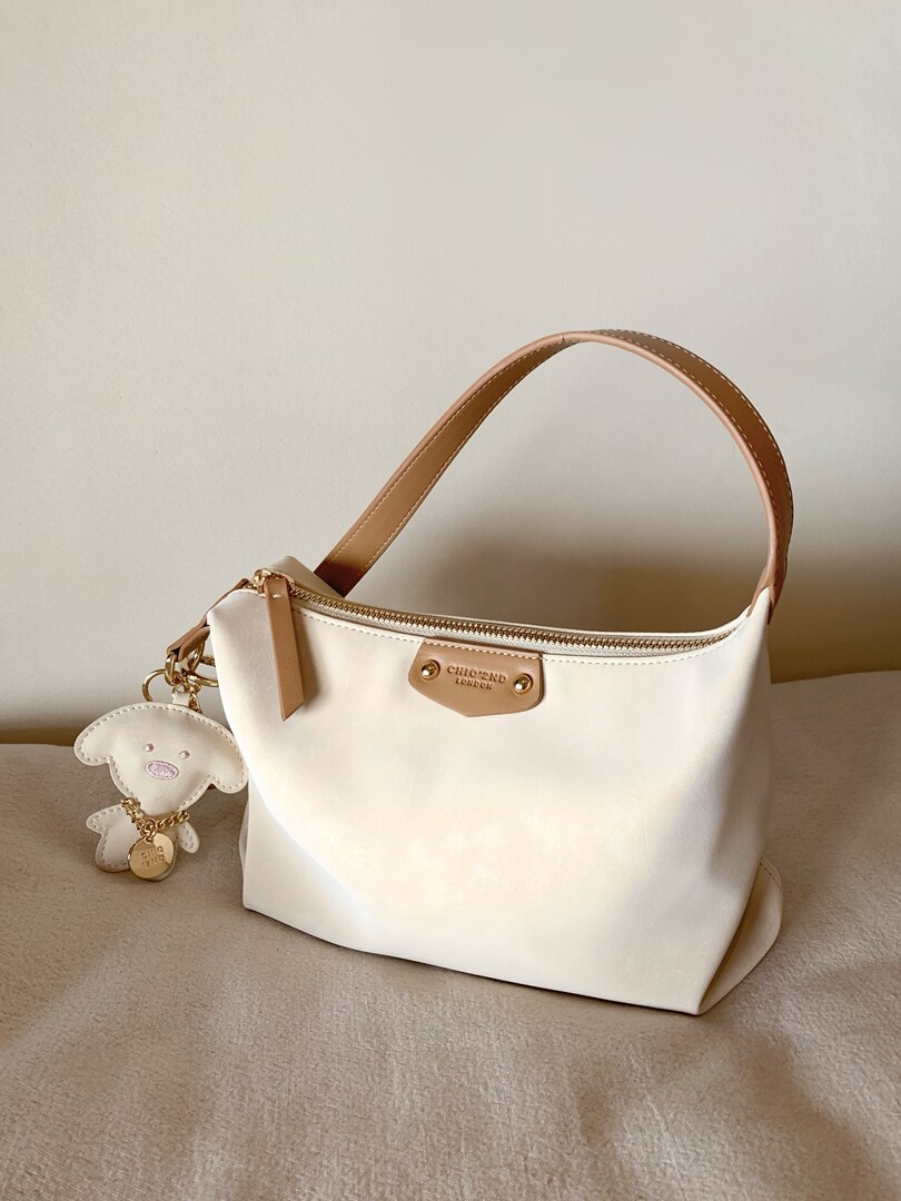 Cream handbag with tan handle and bear charm on a neutral background