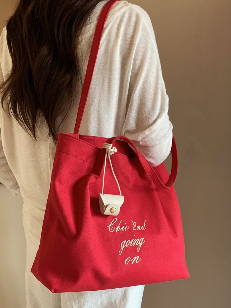 Red canvas tote bag worn over the shoulder with white embroidered text