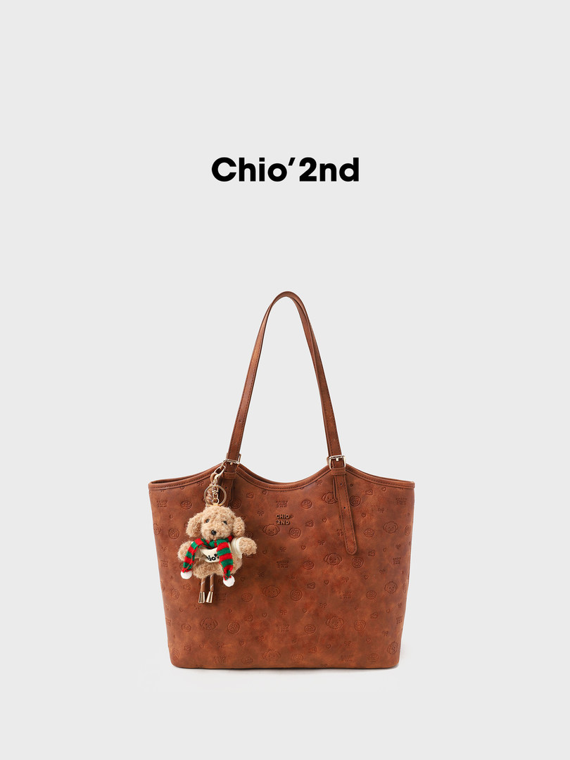Brown tote handbag with a teddy bear charm on a plain background