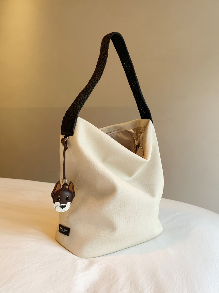 Cream canvas tote bag with black braided handle and dog charm on a bed