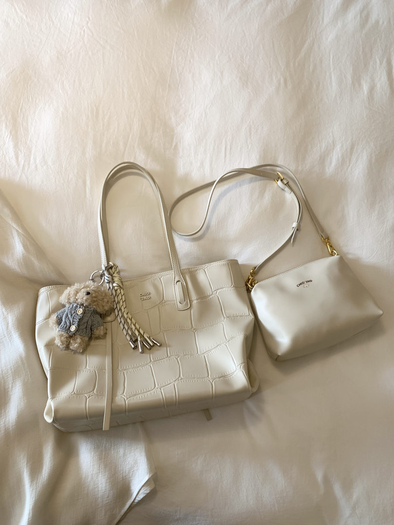 Two cream-colored handbags with a teddy bear charm laid on a fabric background