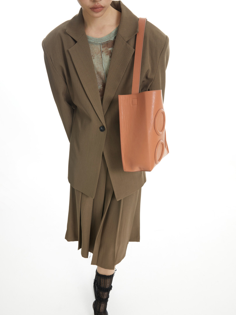 Model wearing an olive blazer and pleated skirt with a tan tote bag