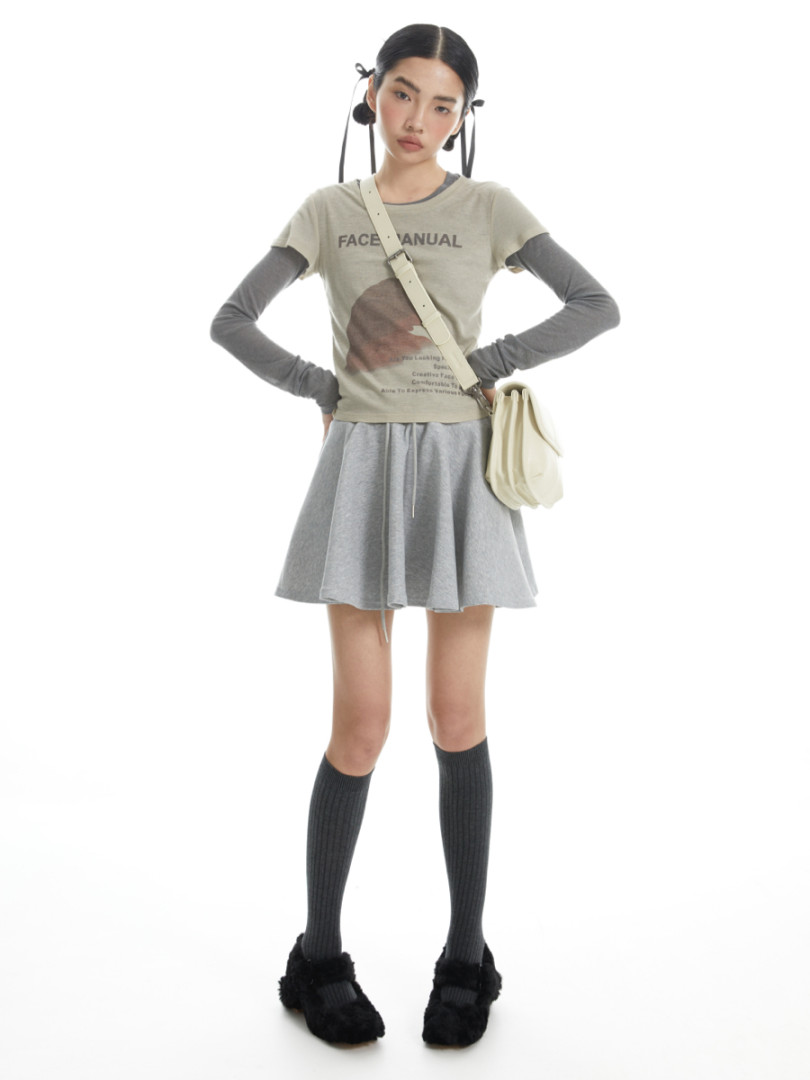 Model wearing a layered casual outfit with graphic tee, gray skirt, and black shoes