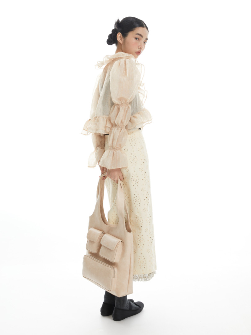 Model wearing a cream sheer ruffled blouse and floral skirt while holding a beige tote bag