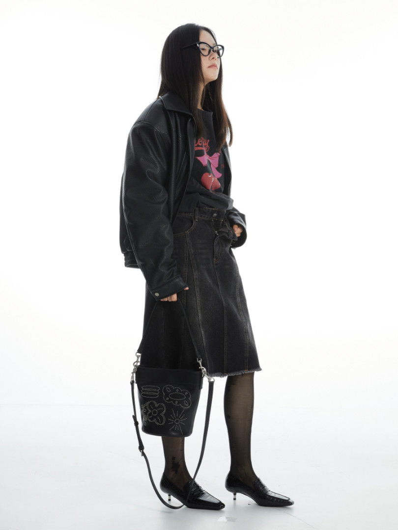 Model wearing a black leather jacket, graphic tee, denim skirt, tights, and heels with a black bucket bag