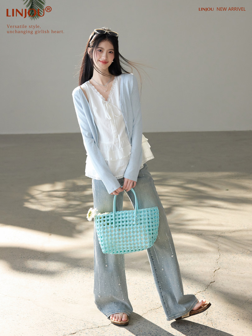 Model wearing a white lace top, light blue cardigan, flared jeans, and holding a woven tote bag.