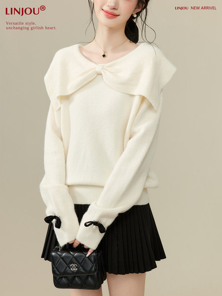 Model wearing a cream bow-detail sweater with black pleated skirt and handbag