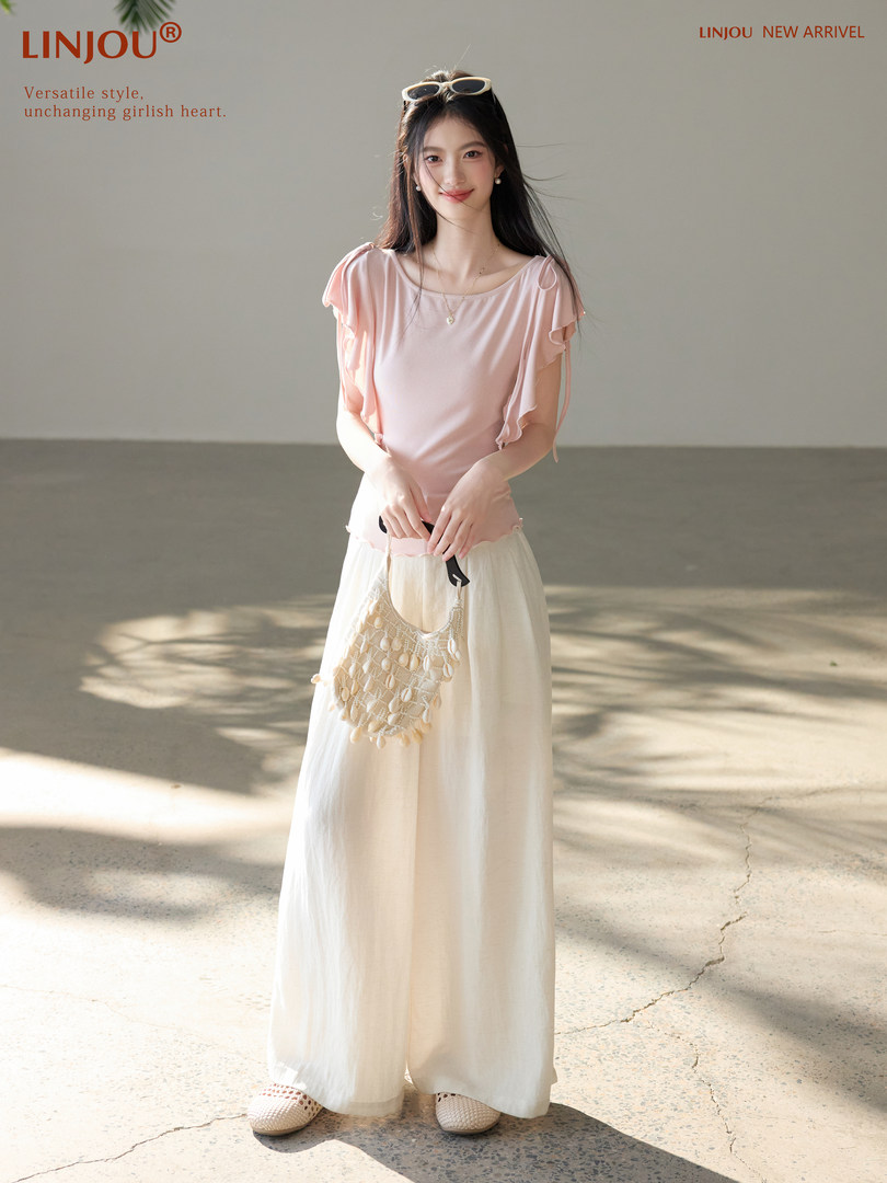 Model wearing a light pink ruffled top with white wide-leg pants and a small shell bag