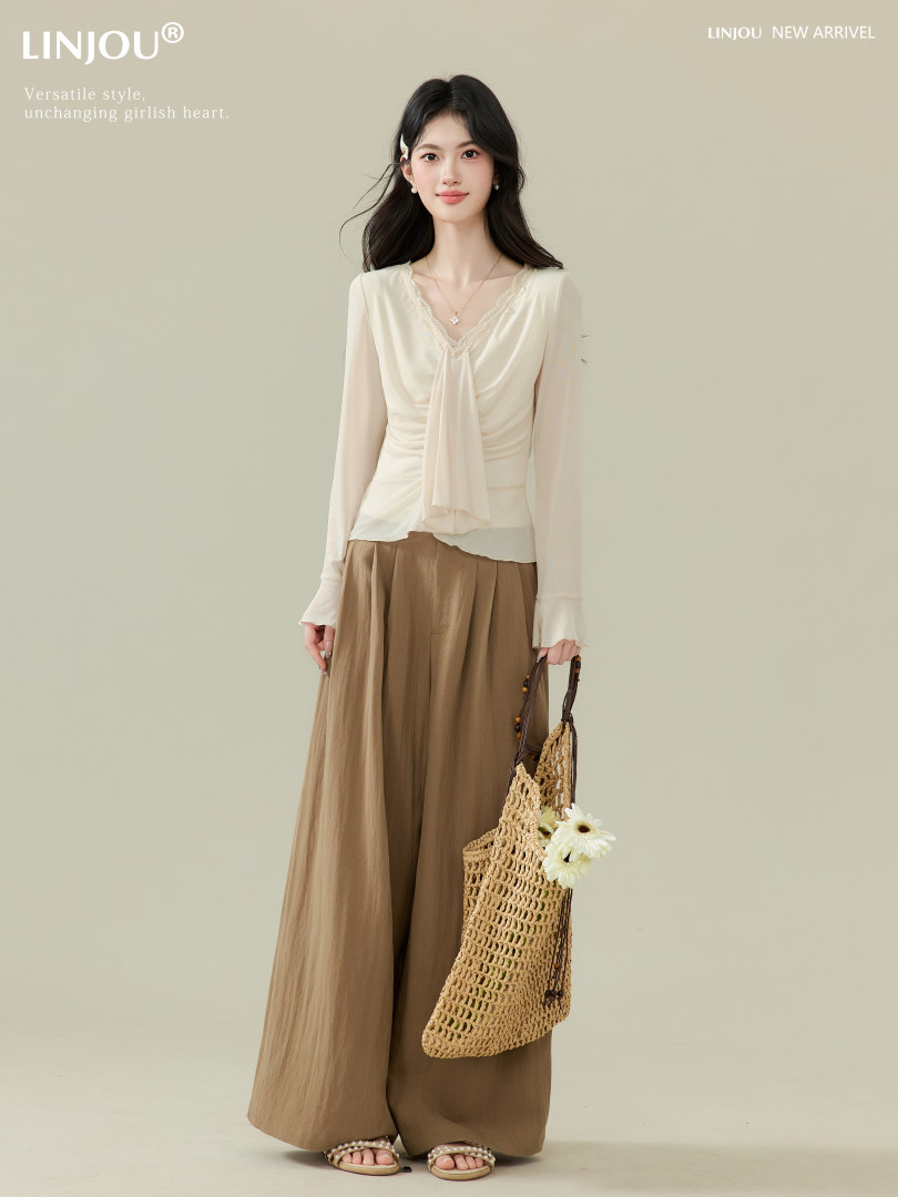Model wearing a cream draped blouse with wide-leg brown pants and a woven tote bag