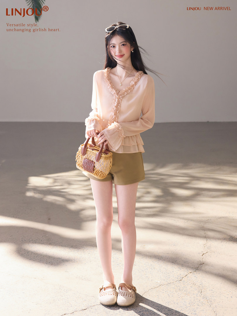 Model wearing a light peach ruffled blouse with shorts and a woven handbag