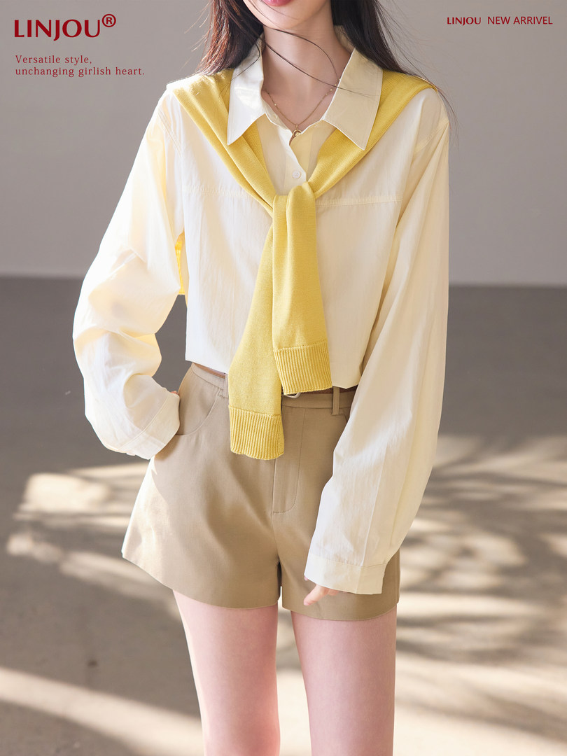 Model wearing a cream oversized shirt with yellow sweater draped over shoulders and beige shorts