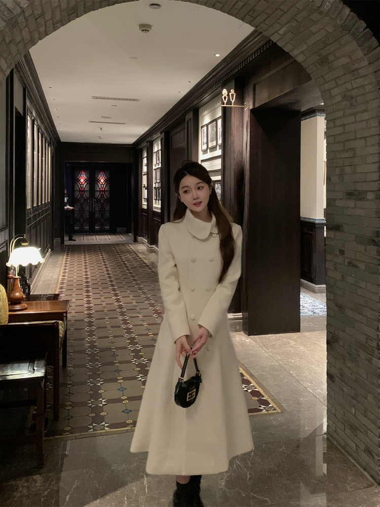 Woman wearing a cream double-breasted long coat in an indoor hallway