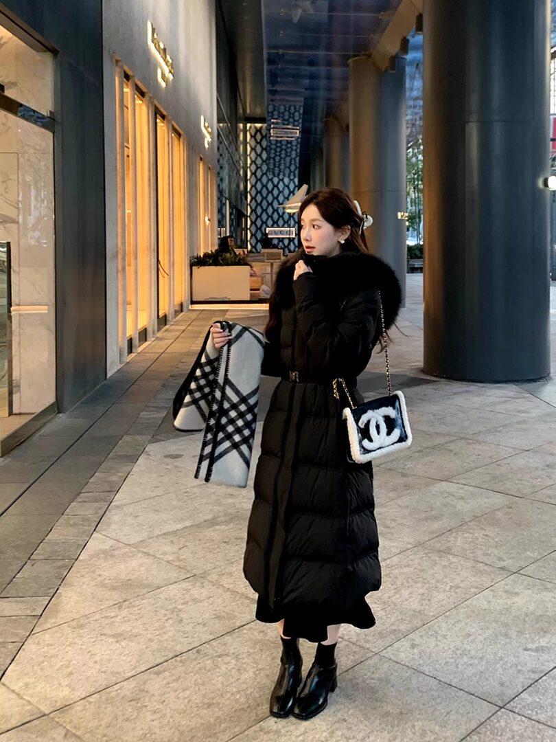 Woman wearing a long black padded winter coat with fur collar and a small chain handbag