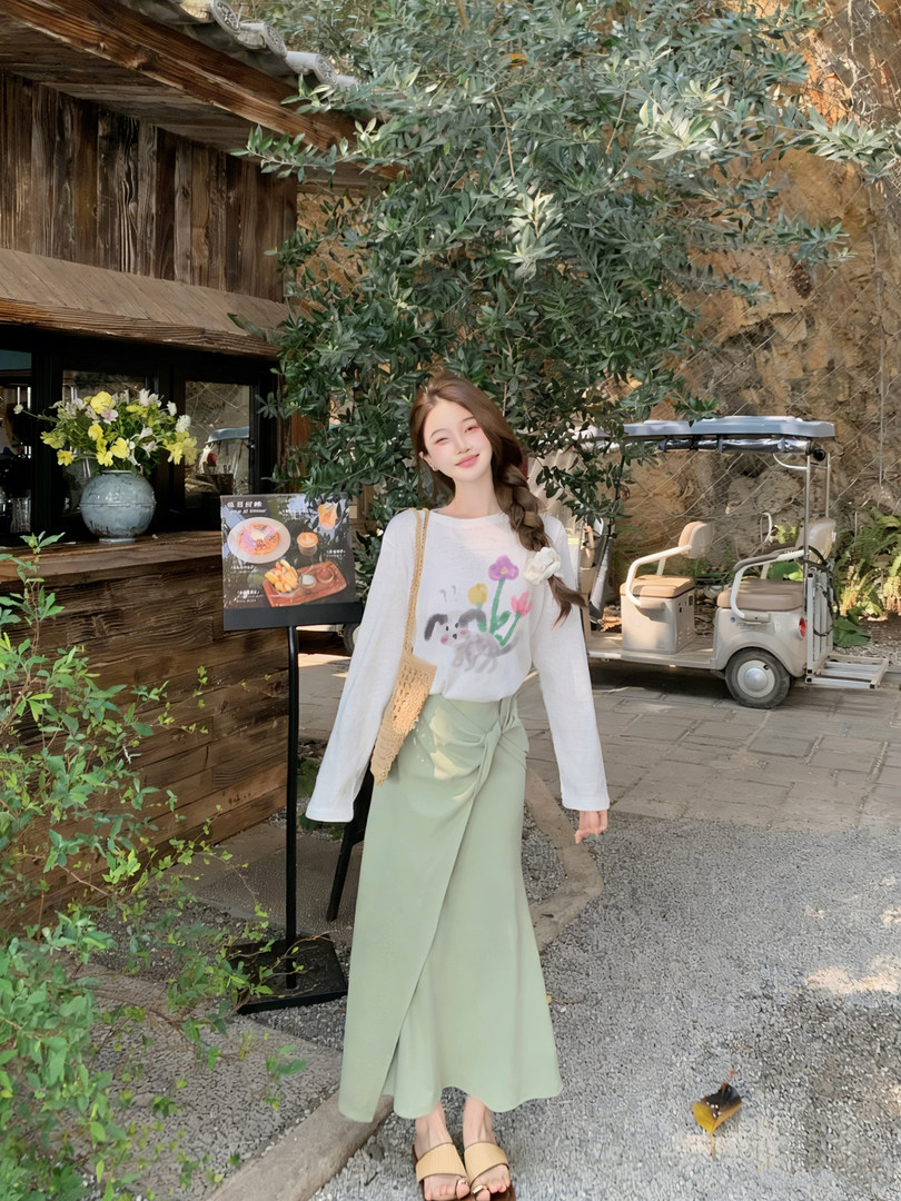 Woman wearing a white graphic long-sleeve top with a mint green wrap skirt and woven shoulder bag outdoors