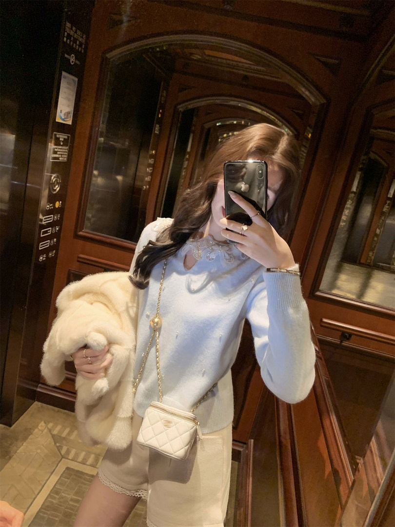 Mirror selfie of a woman wearing a light blue sweater, cream shorts, and a quilted mini crossbody bag