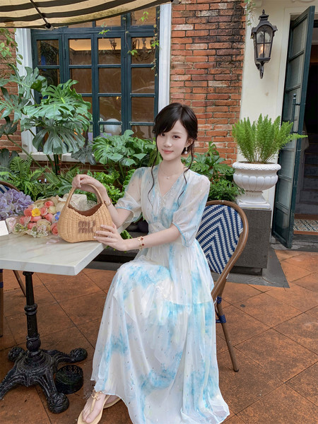 Woman wearing a light blue and white flowy dress while holding a woven handbag outdoors