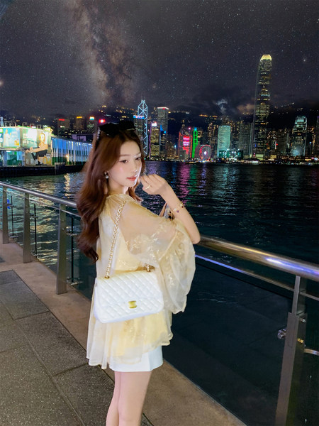 Woman wearing a pale yellow dress with a white quilted handbag at a nighttime waterfront cityscape
