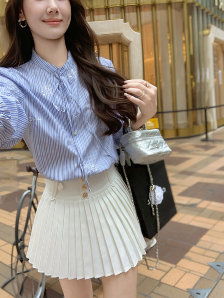 Woman wearing a light blue striped blouse with a white pleated skirt and small handbag