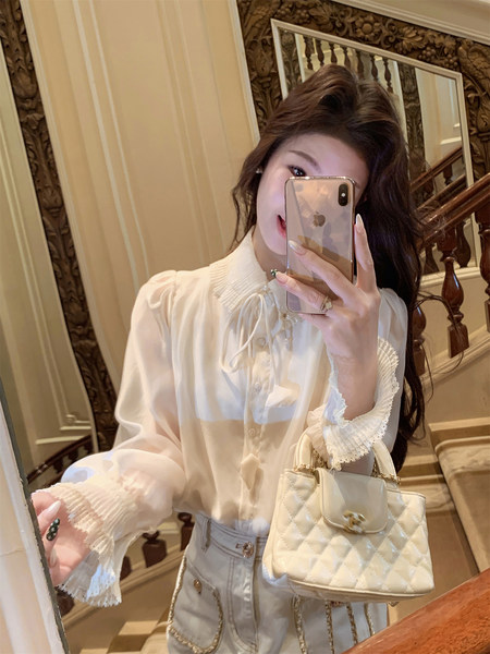 Mirror selfie of a woman wearing a sheer cream blouse and light skirt, holding a quilted cream handbag