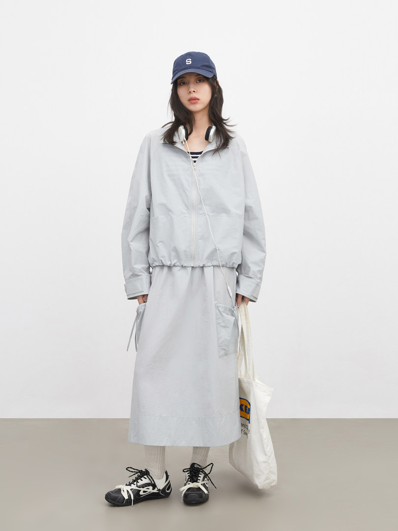 Model wearing a light gray layered outfit with cap, jacket, and long skirt