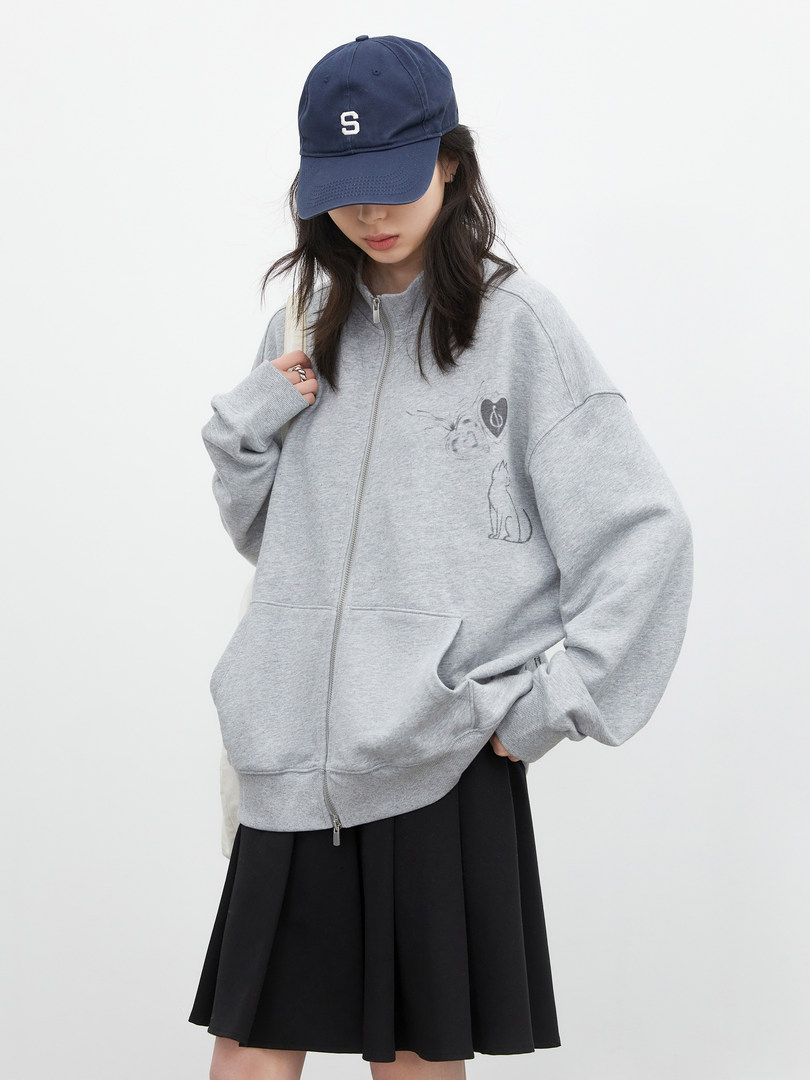 Model wearing a gray zip-up hoodie with graphic details and a navy baseball cap