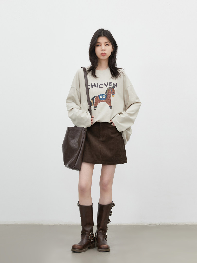 Model wearing a cream graphic sweatshirt with a brown skirt, tall boots, and a dark tote bag