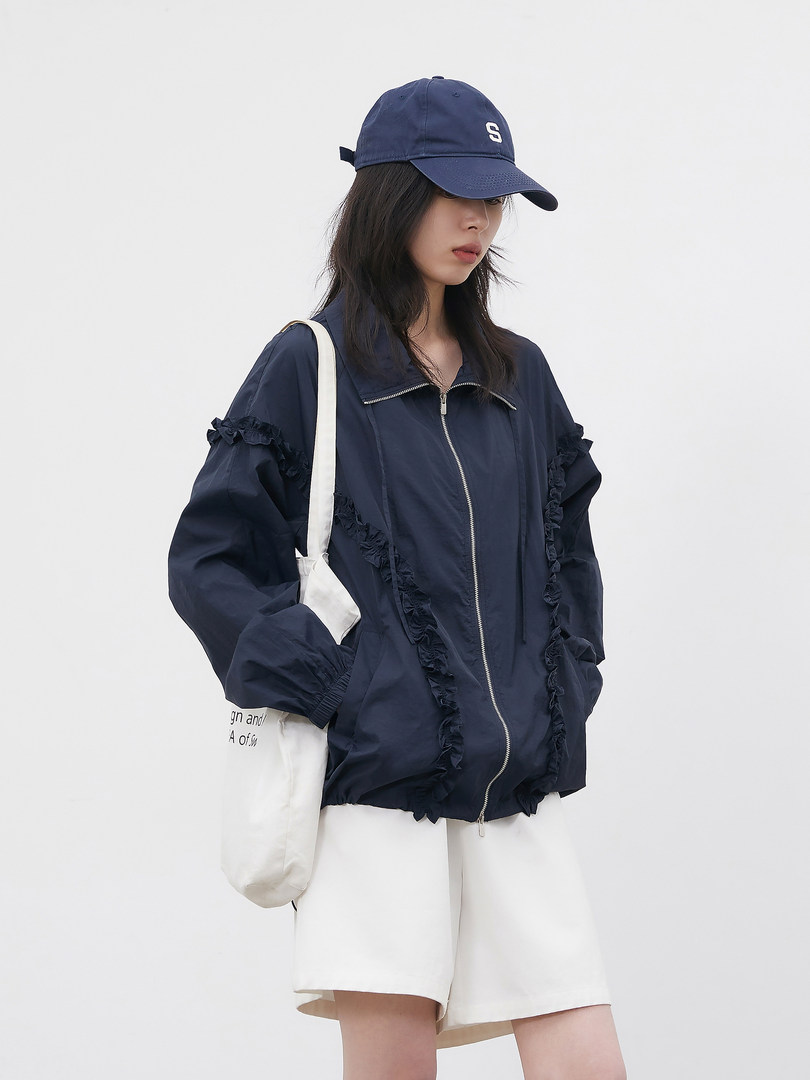 Model wearing a navy zip-up jacket with ruffled details, white shorts, and a baseball cap