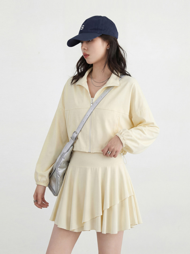 Model wearing a pale yellow cropped jacket and matching skirt with a navy cap and silver crossbody bag