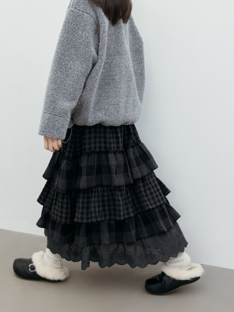 Model wearing a gray fleece pullover with a layered black plaid skirt and fuzzy slip-on shoes