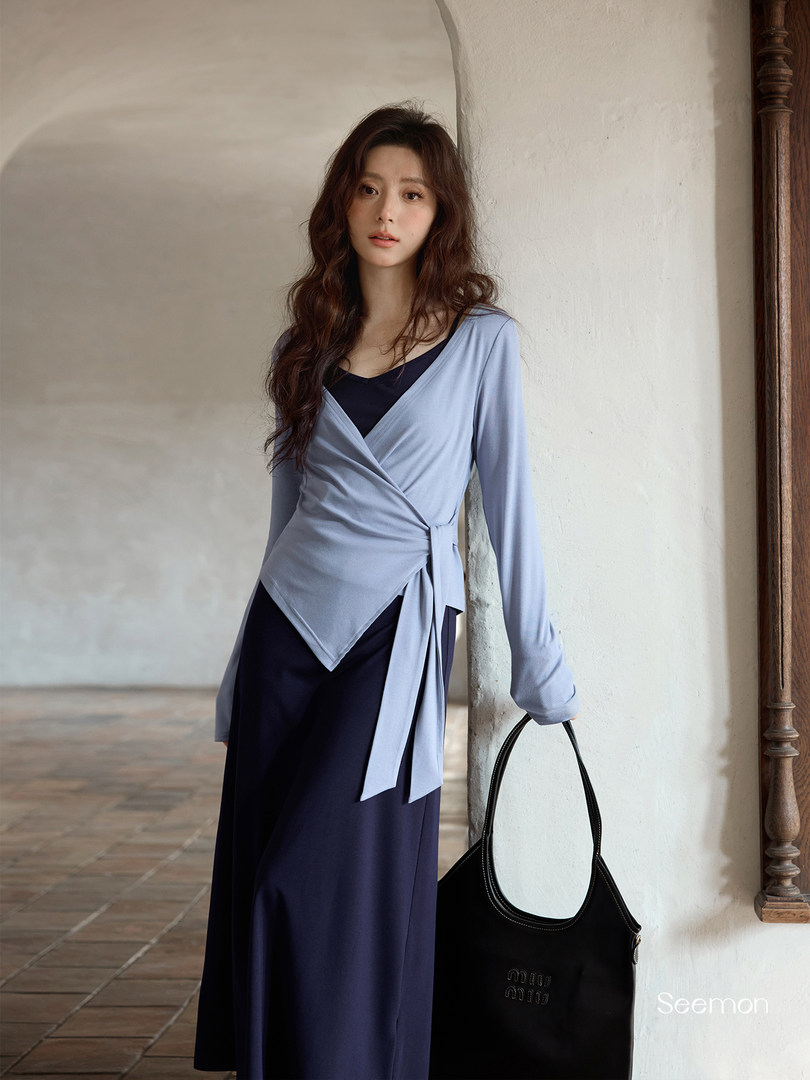 Woman modeling a light blue wrap-style long-sleeve top over a navy dress with a black handbag