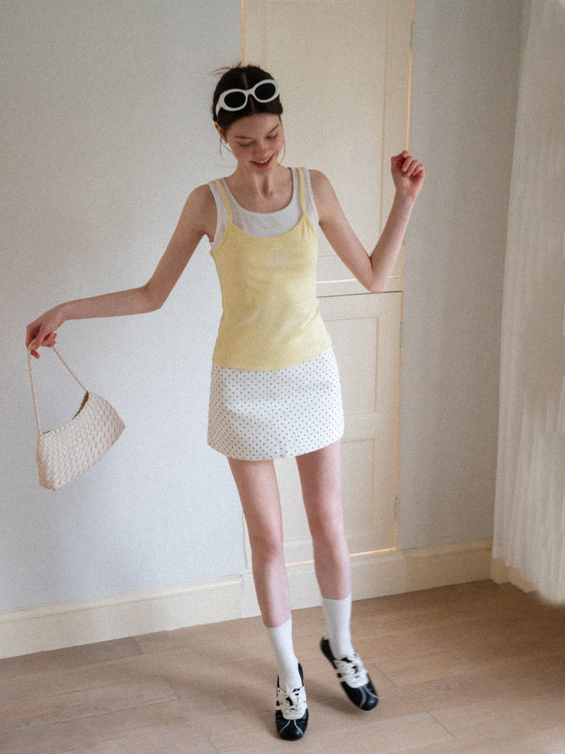 Model wearing a yellow tank top, white mini skirt, and black shoes holding a woven handbag