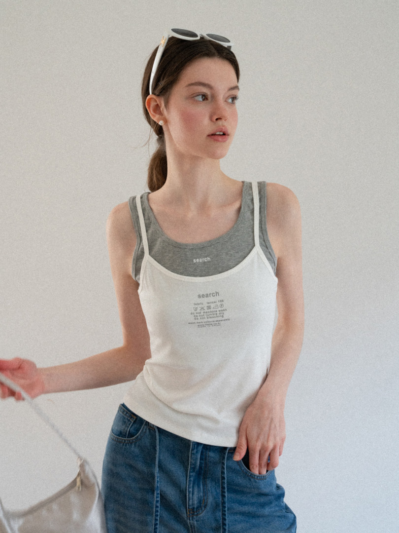 Woman wearing a white and gray layered tank top with printed text and blue jeans