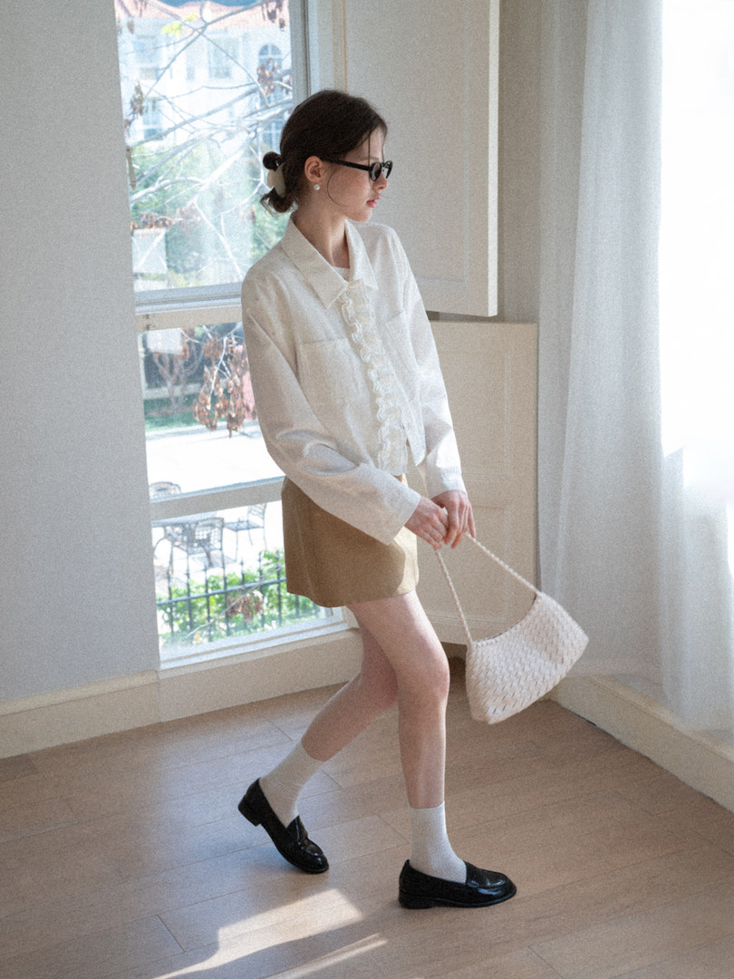 Woman wearing a white ruffled blouse, beige shorts, black loafers, and a woven handbag