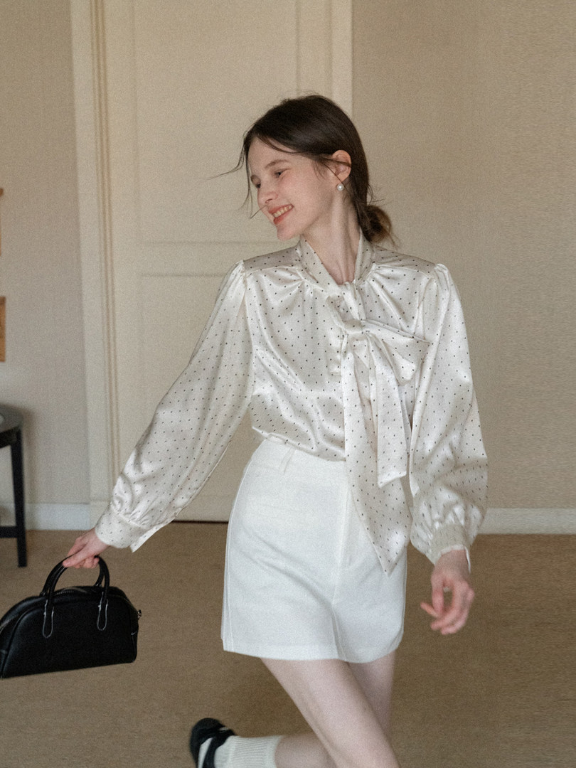 Woman wearing a white satin polka dot bow blouse with white shorts and a black handbag