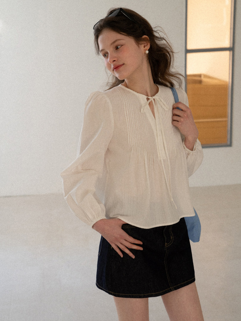 Model wearing a cream sheer long-sleeve blouse with a tie neckline and black denim skirt