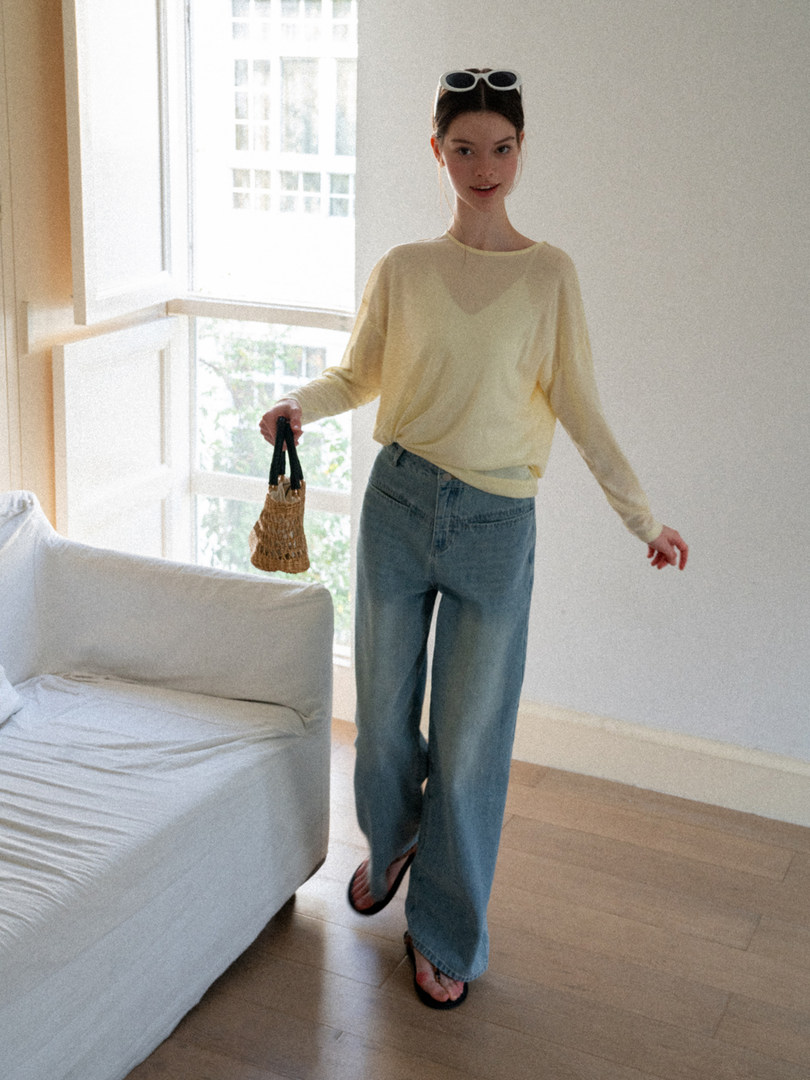 Woman wearing a pale yellow long-sleeve top and blue jeans while holding a small woven bag