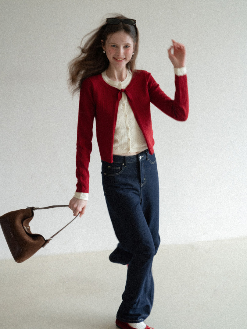 Woman wearing a red cardigan, cream top, and dark jeans while holding a brown handbag