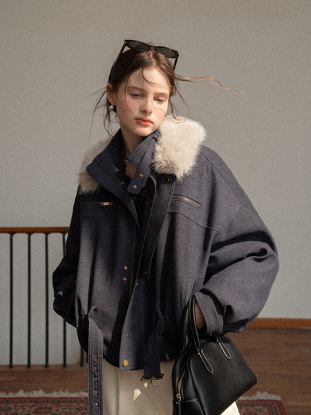 Model wearing a dark oversized coat with a fur collar and carrying a black handbag