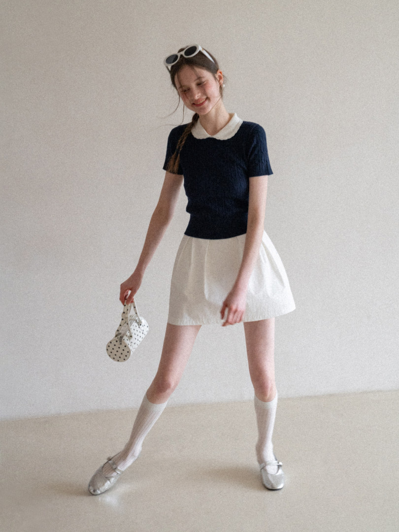 Model wearing a black short-sleeve top with a white collar and a white skirt, styled with socks and flats