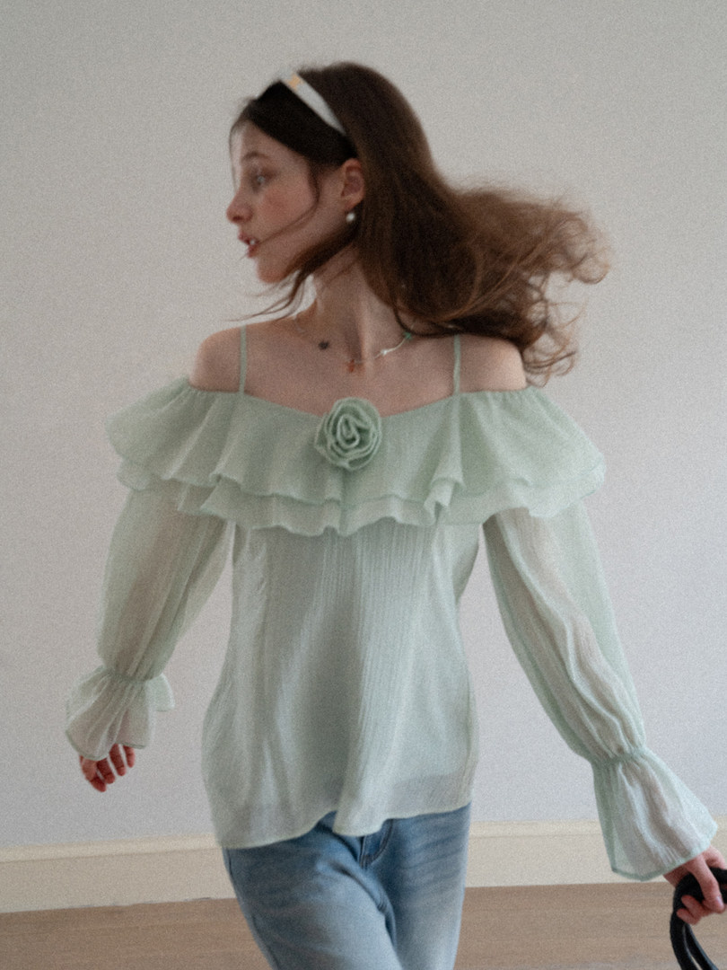 Model wearing a mint green off-shoulder sheer blouse with long sleeves and ruffle detail