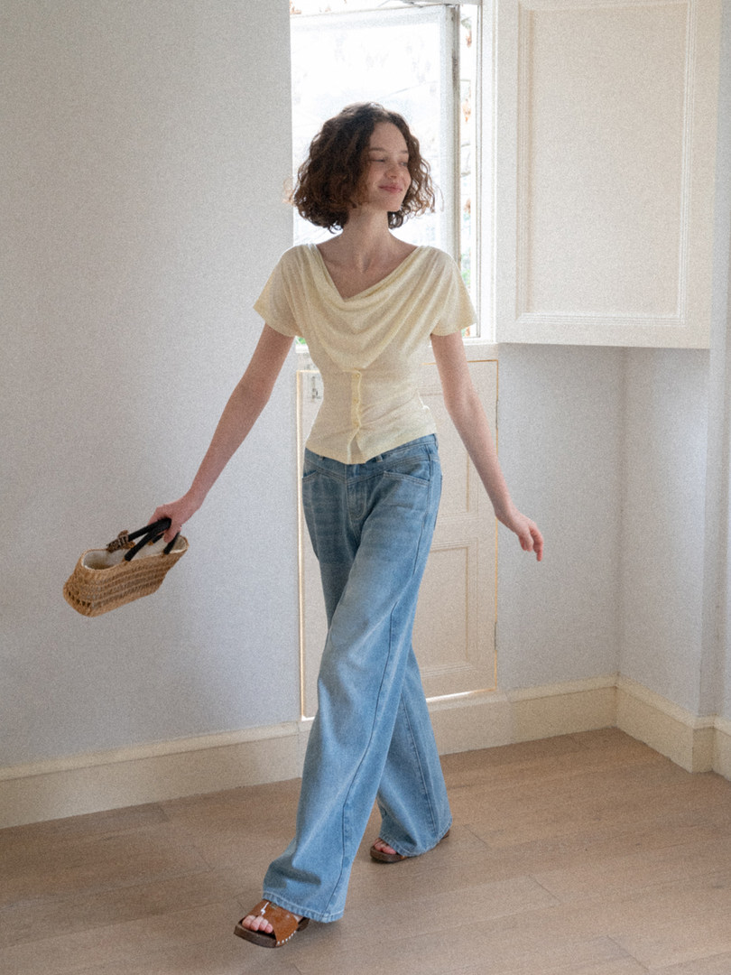 Model wearing a pale yellow draped top with wide-leg blue jeans and holding a woven handbag