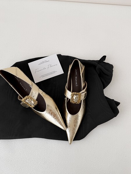 Gold pointed-toe embellished flats displayed on a black fabric backdrop