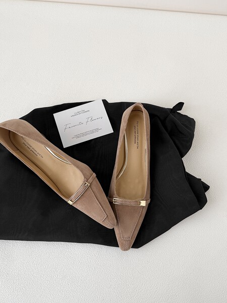 Pair of taupe pointed-toe flat shoes with gold buckle detail laid on a black fabric backdrop